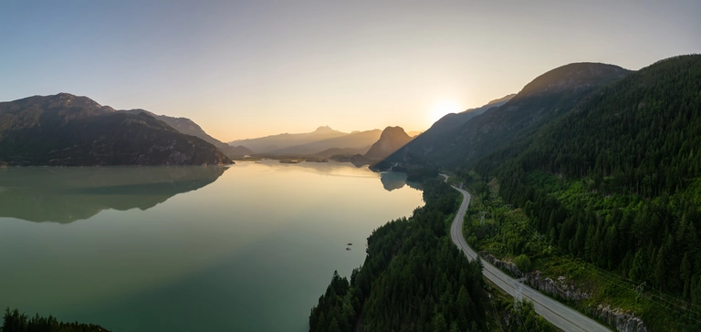 Straße entlang eines malerischen Sees mit grünen Hügeln und Sonnenuntergang im Hintergrund